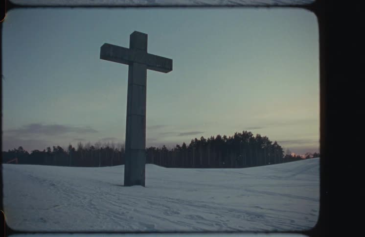 A vintage film shot captures a large stone cross standing alone in a snow-covered field at dusk