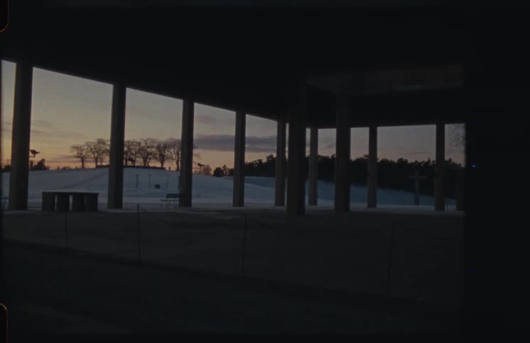 A solemn view from a pillared memorial hall overlooking a snowy cemetery with a large cross at sunset