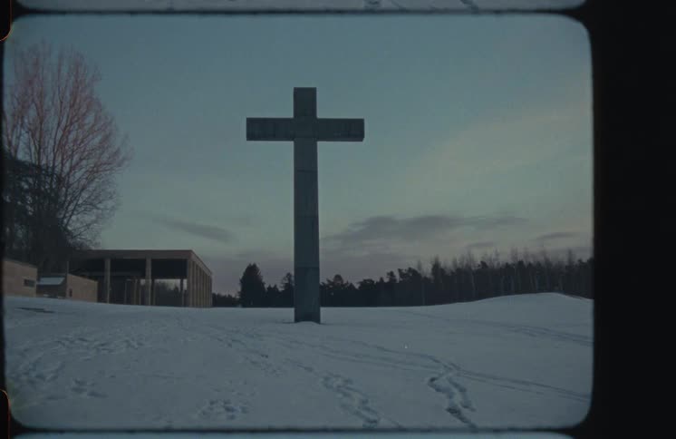 A large stone cross stands in a desolate, snow-covered landscape at dusk, captured on vintage 8mm film