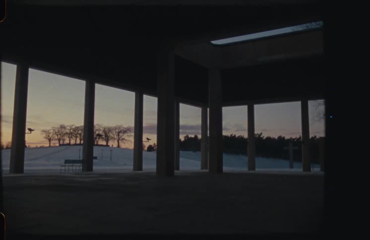 A serene sunset view of a snowy cemetery with a cross, seen from inside a dark, modern, pillared structure on vintage film