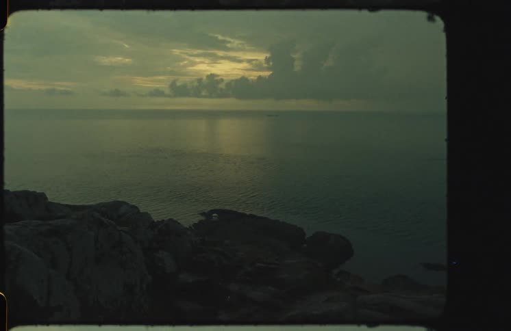 A serene, moody seascape is captured on vintage film, showing the calm ocean from a rocky cliff at dusk