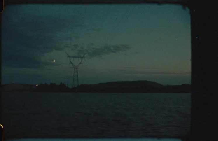 A vintage 8mm film captures an electrical pylon silhouetted against the night sky over a still lake, with a crescent moon peeking through the clouds