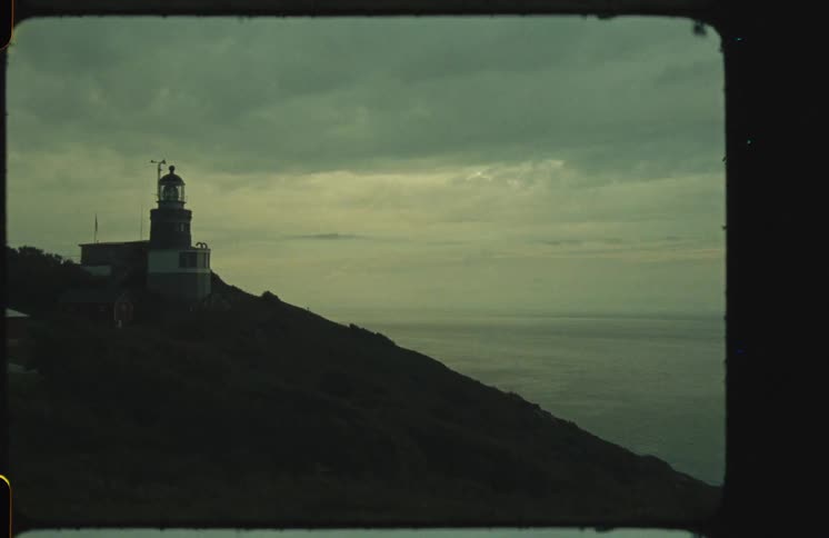 Vintage 8mm footage captures a moody scene of a lighthouse on a remote hill overlooking the ocean under a cloudy sky