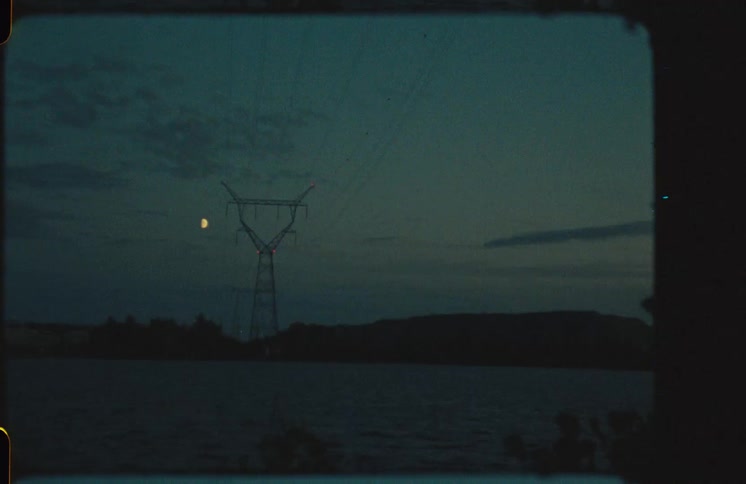 Vintage Super 8 footage shows a half-moon and an electricity pylon silhouetted against the twilight sky over a calm lake