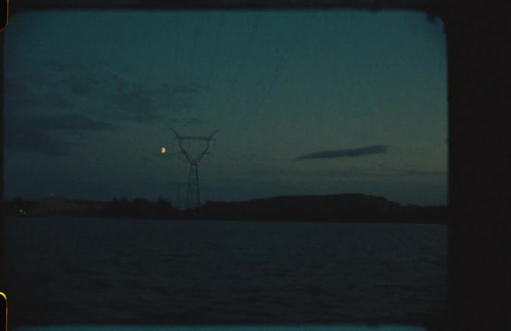 Vintage 8mm film captures a lonely electrical tower and a crescent moon over dark water at twilight