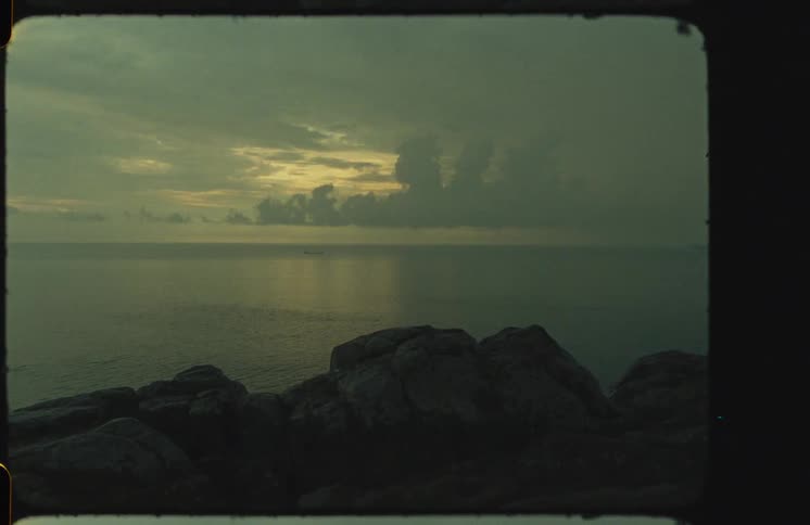 A moody, vintage film shot captures a calm ocean at dusk from a rocky shore under a cloudy sky
