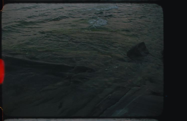 Vintage 8mm film of dark water rippling against a rocky shore with light leaks