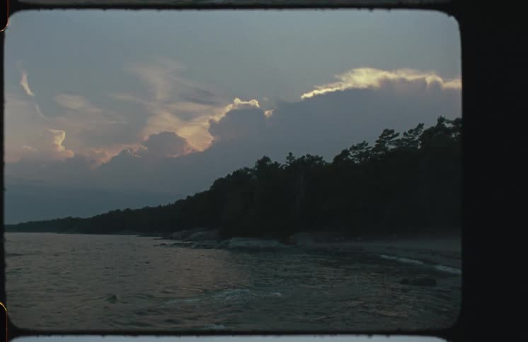 Vintage film captures a moody coastline at dusk, with gentle waves lapping against the shore and dramatic clouds in the sky