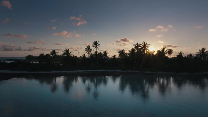 A serene sunset casts a golden glow over a silhouetted tropical island, its palm trees perfectly reflected in the still, glassy water