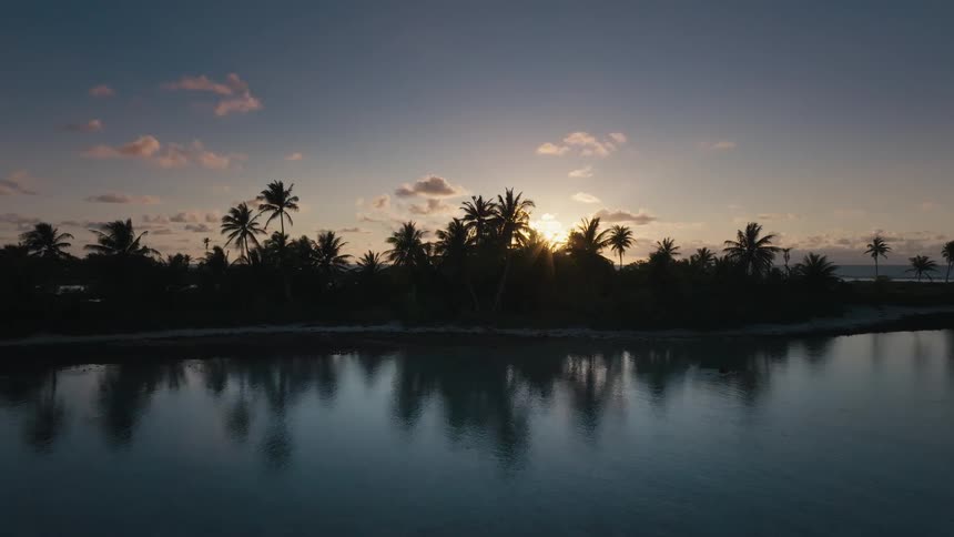A peaceful tropical island is silhouetted by the setting sun, its palm trees perfectly reflected in the calm foreground water