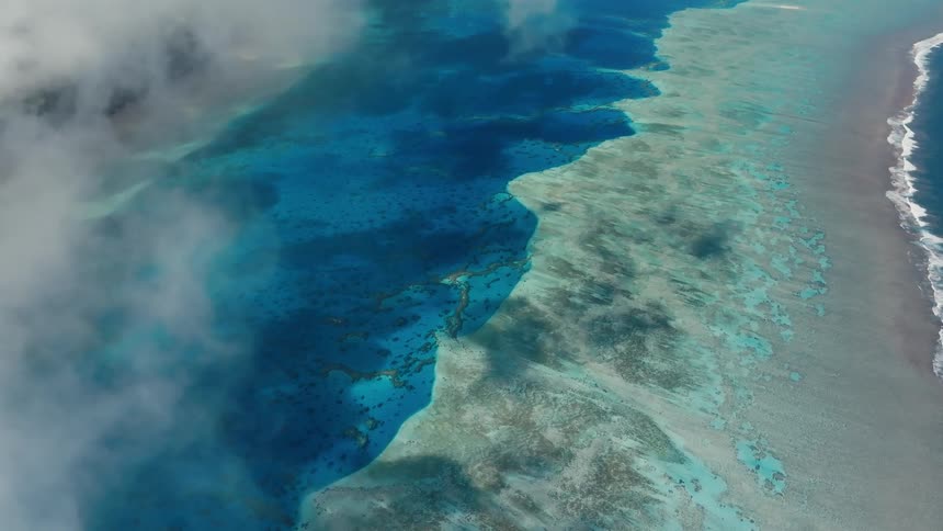 A serene aerial view from above the clouds reveals a stunning turquoise lagoon and coral reef system