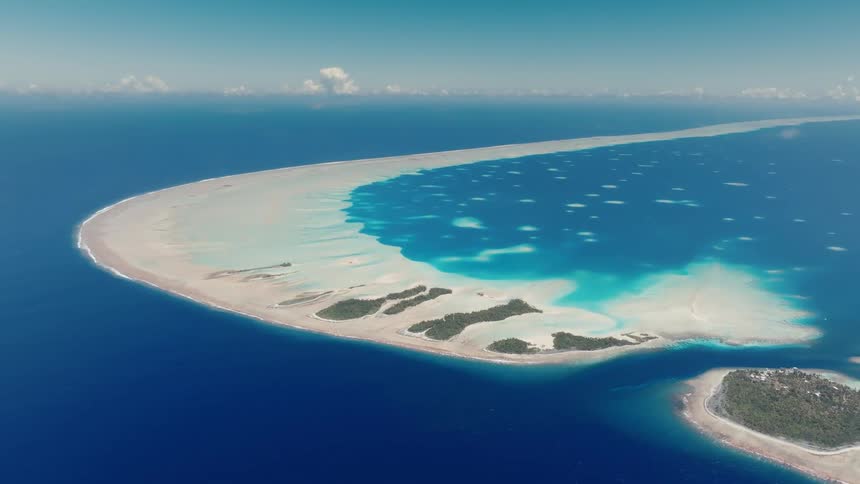 A breathtaking aerial perspective reveals a curved tropical atoll with a turquoise lagoon, white sands, and lush islets surrounded by the deep blue Pacific Ocean