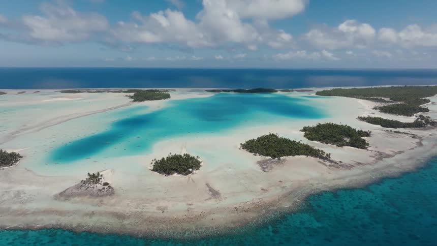 A stunning static aerial shot captures the breathtaking beauty of a tropical atoll with turquoise lagoons and small palm-covered islands in the middle of the vast blue ocean