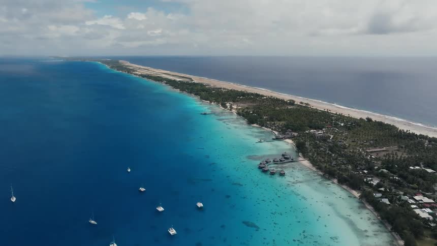 A breathtaking aerial view showcases a long tropical atoll with sailboats resting in the calm, vibrant turquoise lagoon