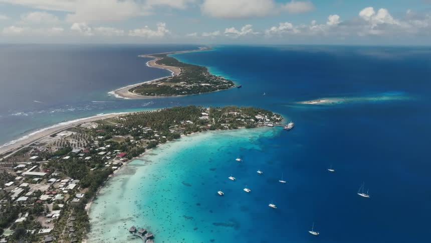 An expansive aerial view shows a stunning tropical atoll with a turquoise lagoon, deep blue ocean, and anchored sailboats