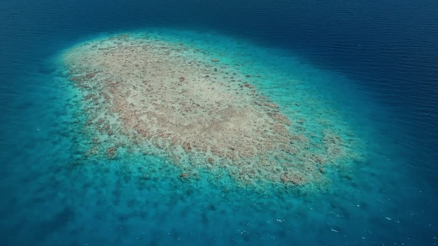 A serene aerial view shows a stunning coral atoll emerging from the deep blue sea, surrounded by crystal-clear turquoise waters