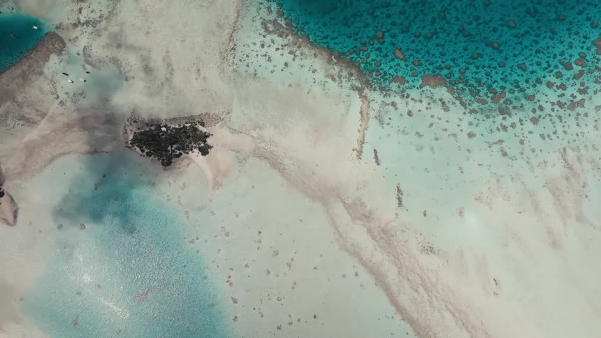 An ascending top-down aerial shot reveals a stunning tropical paradise with turquoise lagoons, white sandbanks, and a vibrant coral reef surrounding a small, isolated islet