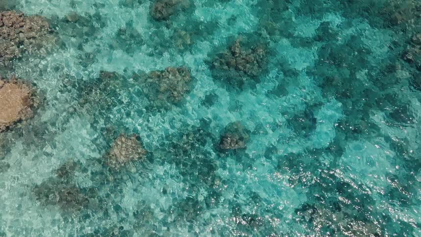 An aerial top-down perspective of the shimmering, crystal-clear turquoise sea with visible coral formations