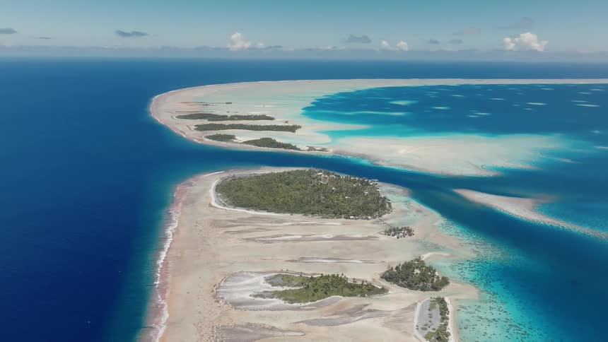 A stunning high-angle aerial view shows a tropical atoll with lush green islands surrounded by the deep blue ocean and a vibrant turquoise lagoon