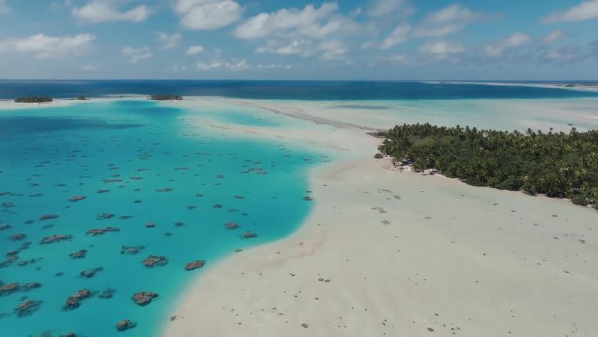 A breathtaking static aerial shot captures the vibrant turquoise waters and white sand of a remote tropical atoll under a blue sky with fluffy clouds