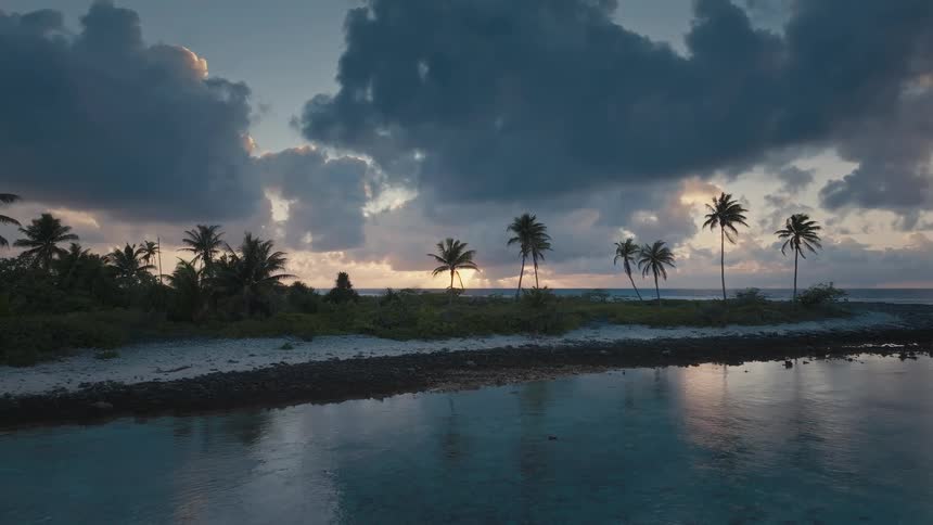 Slow aerial pan revealing a serene tropical island with palm trees silhouetted against a dramatic cloudy sunrise over the ocean