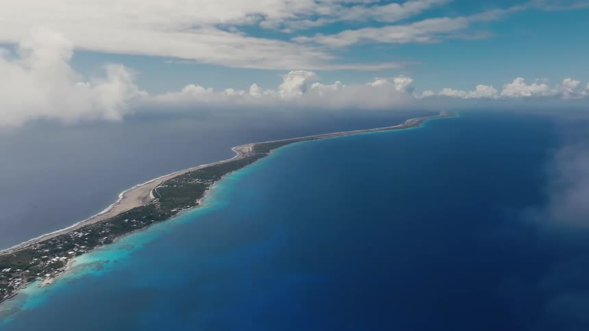 A serene and expansive aerial cinemagraph captures a long, narrow tropical atoll stretching across the vast, deep blue ocean