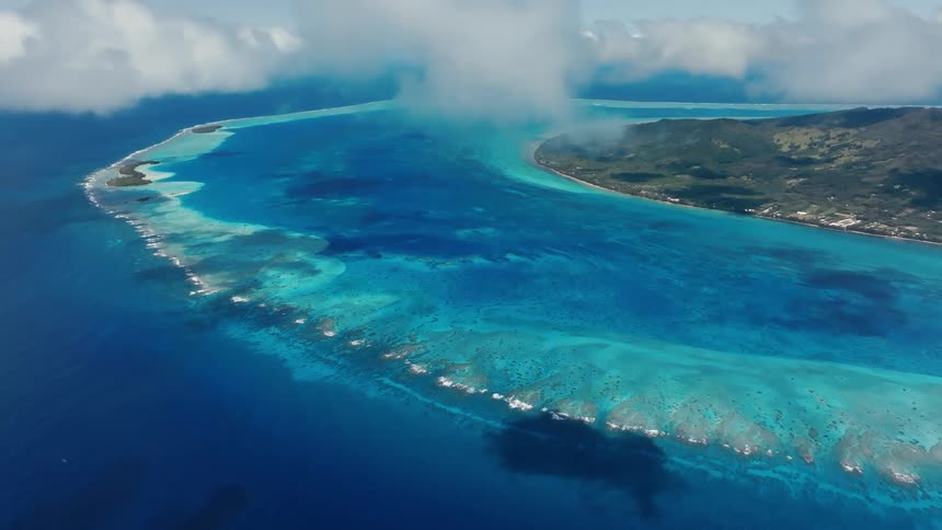 An epic aerial cinemagraph reveals a tropical island paradise with a vibrant coral reef and stunning turquoise lagoon under fluffy clouds