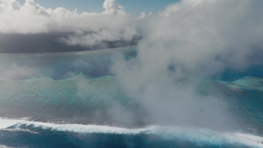A serene aerial view from above the clouds reveals the turquoise waters of a tropical lagoon and breaking waves on a coral reef