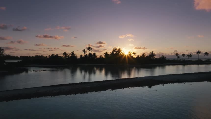 Aerial view slowly pushes over a calm tropical lagoon towards a silhouetted island with palm trees at sunset