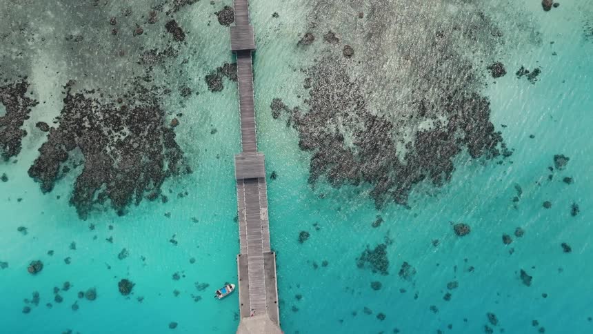 A stunning aerial top-down view slowly ascends over a long wooden pier as a couple begins to kayak in the tranquil, clear turquoise ocean over a coral reef