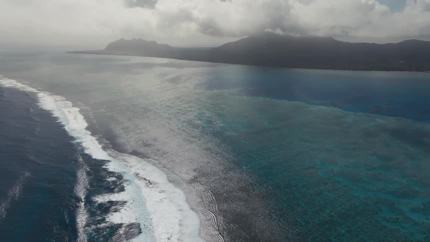 Breathtaking aerial view of ocean waves breaking on a coral reef, separating the deep blue sea from a tranquil turquoise lagoon with a mountainous island in the background