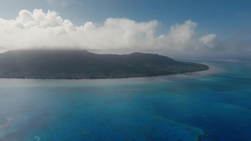 Static aerial view of a majestic tropical island partially covered in clouds, surrounded by the clear turquoise ocean