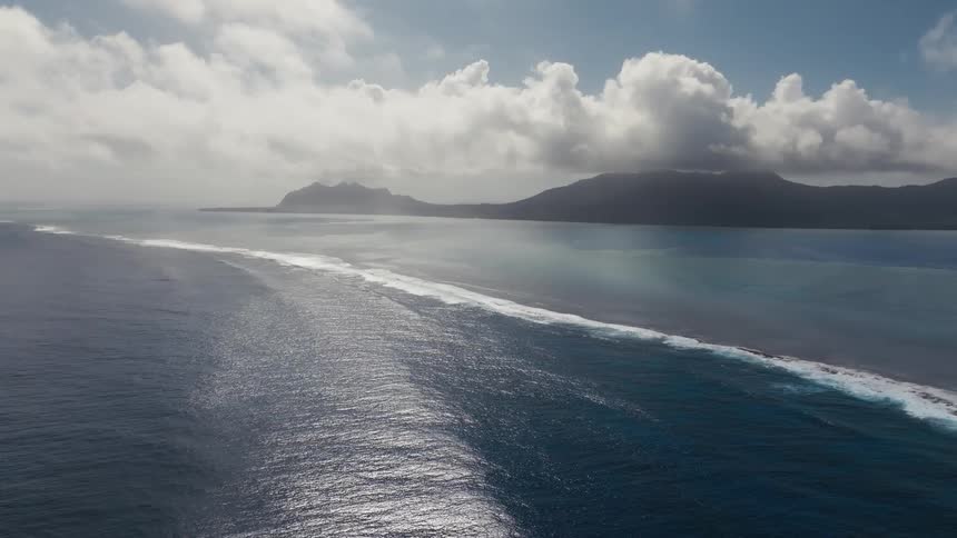 A breathtaking static aerial view captures the vastness of the ocean as sunlight shines on waves breaking along a reef, with a distant tropical island on the horizon