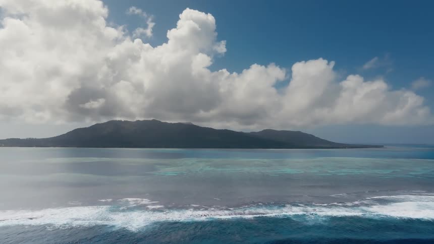 An aerial cinemagraph captures the serene beauty of a tropical island, with ocean waves breaking over a reef under a partly cloudy sky