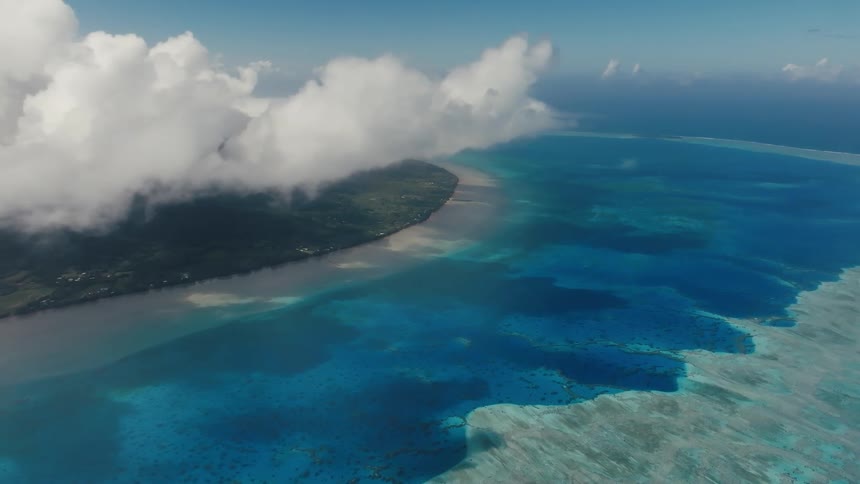 An expansive aerial view of a tropical island and a vast coral reef lagoon under a partially cloudy blue sky