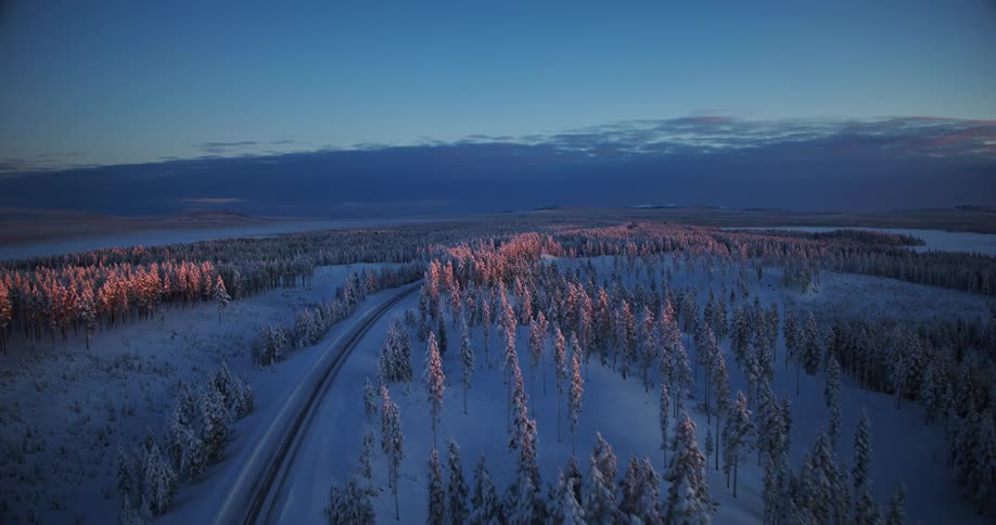 Aerial time-lapse shows a car's light trails on a road through a snowy forest at sunset, with the last light catching the treetops