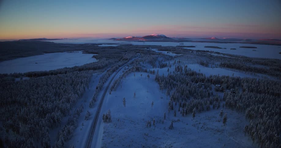 A stunning aerial view shows a snow-covered road running through a vast frozen forest landscape at dusk, with sunlit mountains in the distance