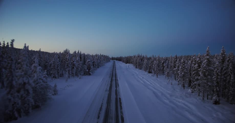 An aerial view descends onto a long, remote road cutting through a vast, snow-covered forest at dusk