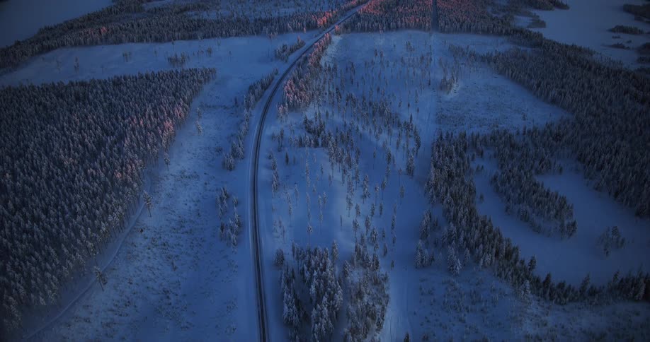 An expansive aerial view reveals a solitary road cutting through a vast, snow-covered forest as the rising sun casts a warm glow on the landscape