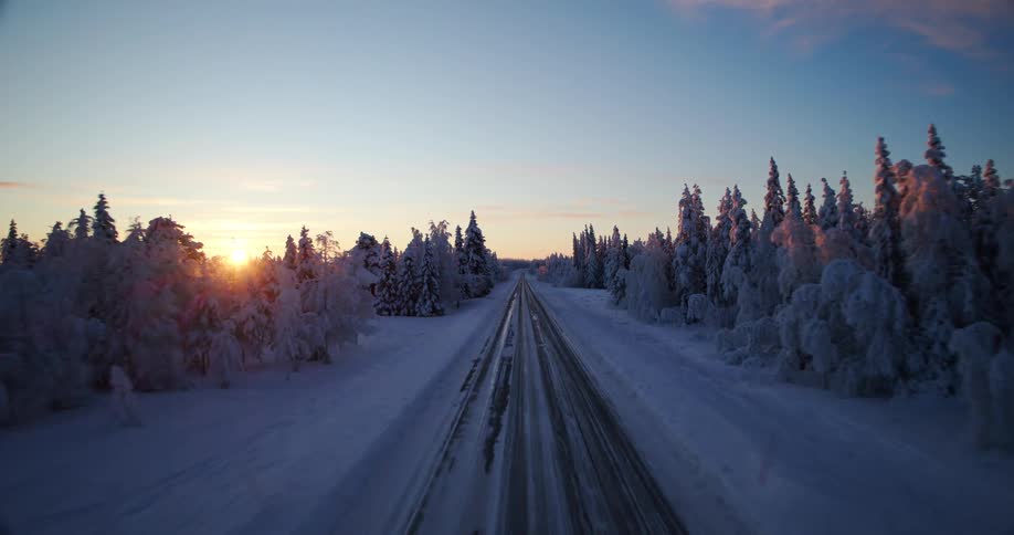 Aerial drone view pulls back over a long snowy road through a frozen pine forest during a beautiful winter sunset