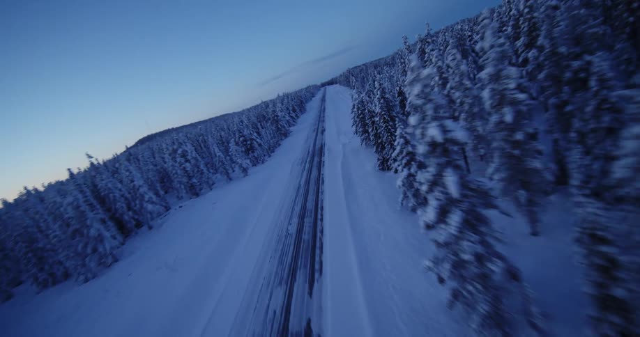 An FPV drone soars over a vast, snow-covered forest before diving down to race along a remote road at twilight