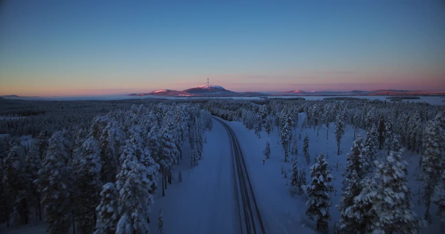 A breathtaking aerial shot follows a snowy road through a winter forest landscape during the twilight hours