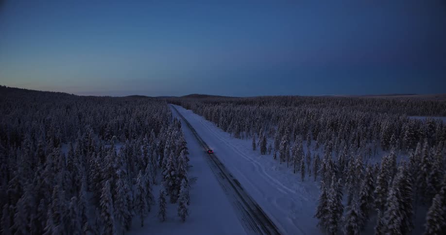 Aerial view of a red car driving through a vast, snow-covered forest in Lapland during the blue hour twilight