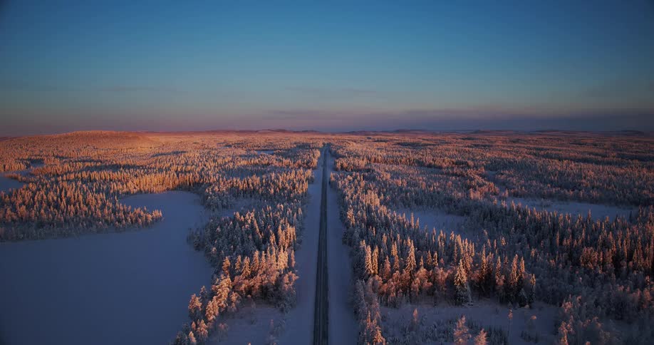 A serene aerial view follows an empty road through a vast, snow-covered Lapland forest, illuminated by the golden glow of a winter sunrise