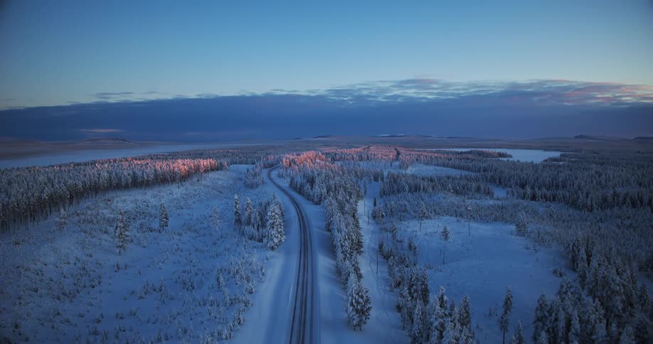 A serene aerial view follows a remote road through a vast, snow-covered forest as a lone car drives towards a beautiful winter sunrise