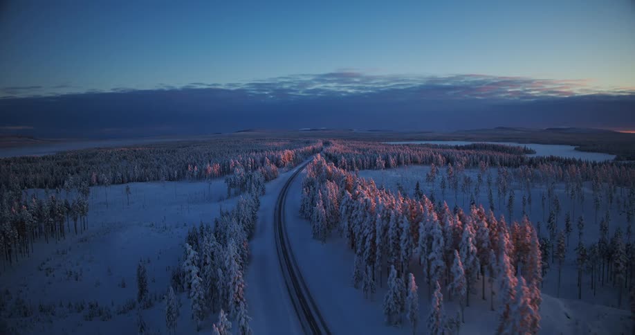 A majestic aerial view of a road winding through a vast, snow-covered forest at sunrise, with a warm glow on the treetops