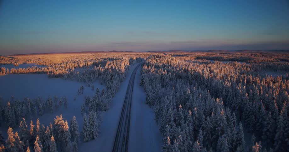 A breathtaking aerial view follows a snowy road through a vast, sun-kissed evergreen forest during a serene winter sunset