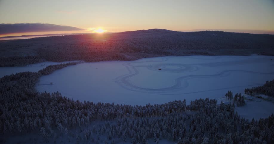 A stunning aerial view reveals a vast winter landscape with a frozen lake and car track as the sun sets over the snow-covered forest
