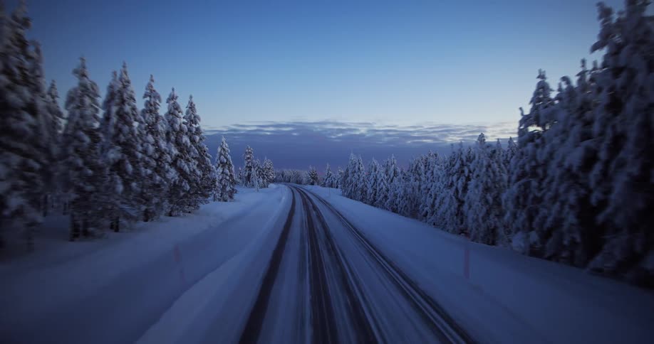 An aerial backwards dolly shot reveals a snow-covered road disappearing into a serene, frozen pine forest during the blue hour