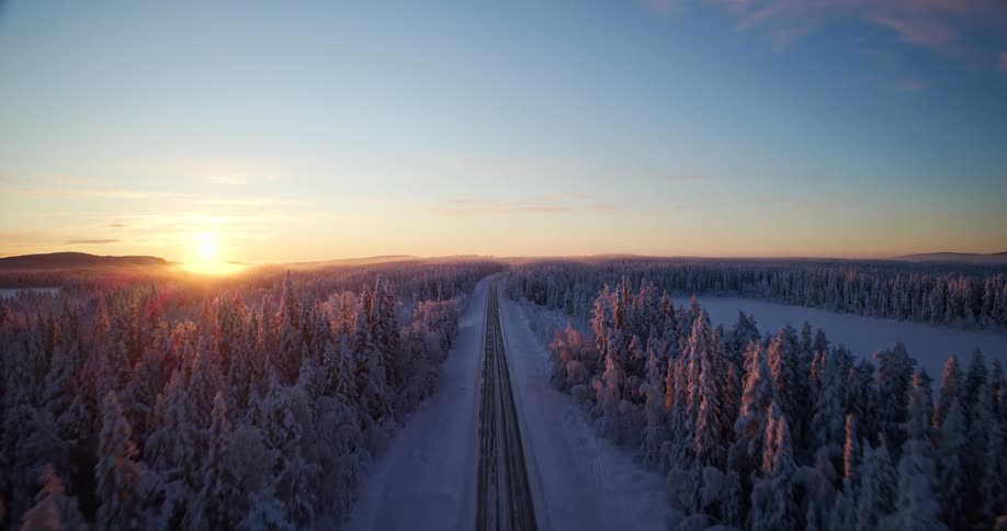 Aerial view pulls back from a snowy road to reveal a vast, frozen forest in Lapland during a stunning golden sunset